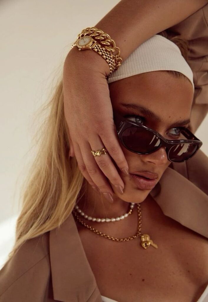 Fashionable woman with sunglasses, gold jewelry, and headband poses stylishly against a soft background.