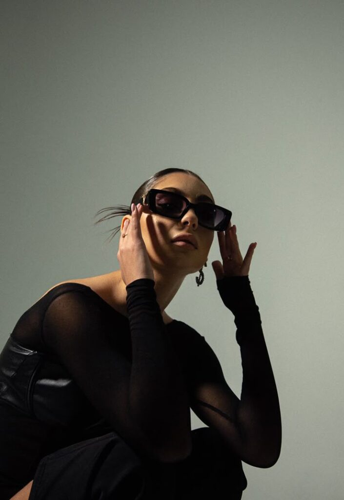 Woman in black outfit and sunglasses posing with hands near face against gray background.
