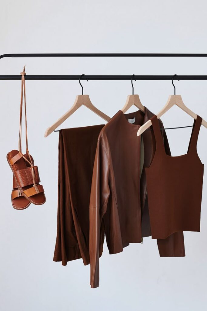 Brown leather jacket, tank top, and pants on hangers; brown sandals hanging on a rack against white background.