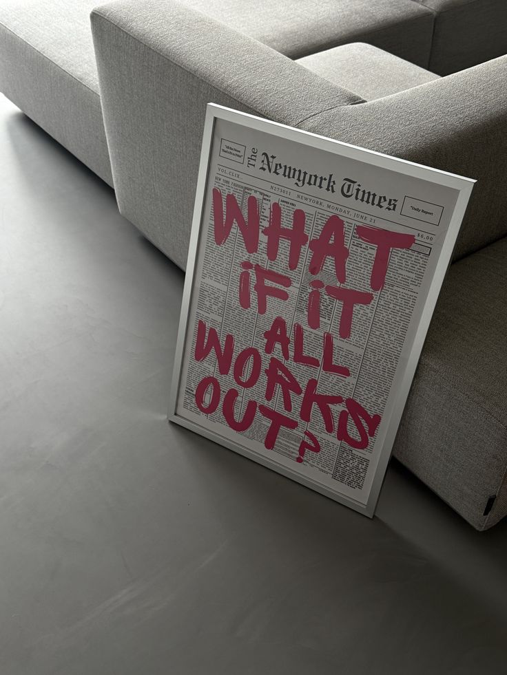 Framed newspaper art with What if it all works out? in red, leaning against a gray sofa on a modern floor.