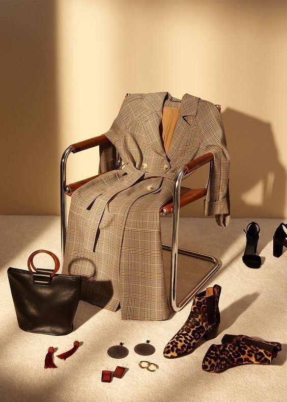 Plaid coat draped over chair with leopard boots, black bag, earrings, and heels on beige carpet in warm lighting.