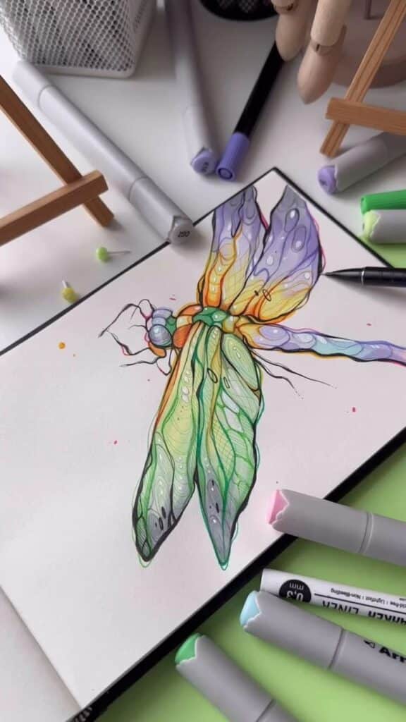 Colorful dragonfly drawing with markers on sketchbook, surrounded by art supplies.