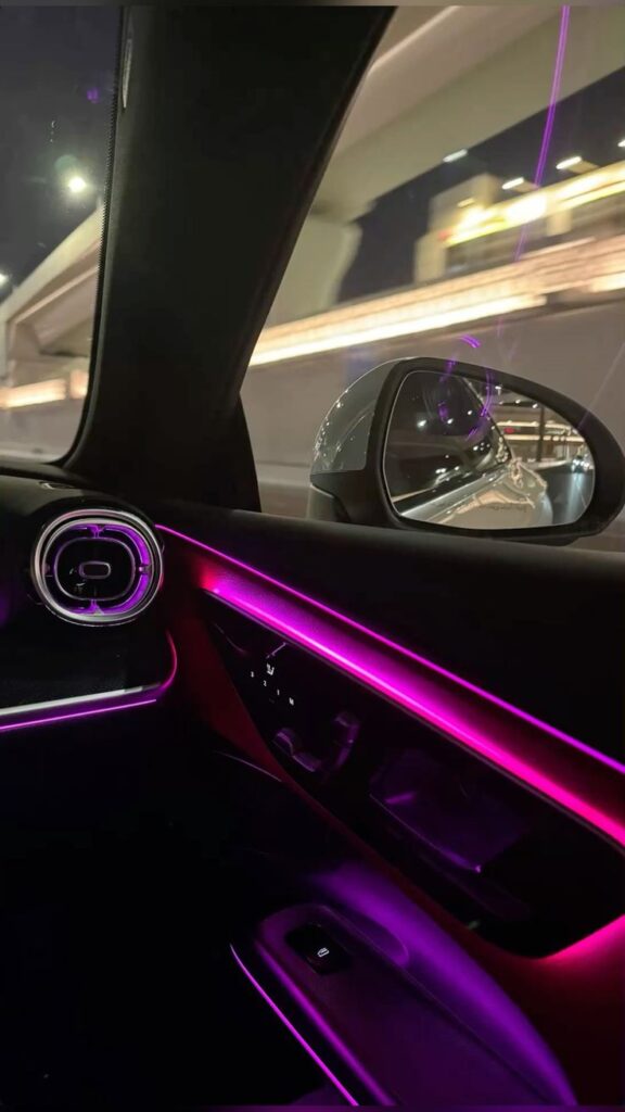 Car interior with neon pink ambient lighting, view of side mirror reflecting highway at night.