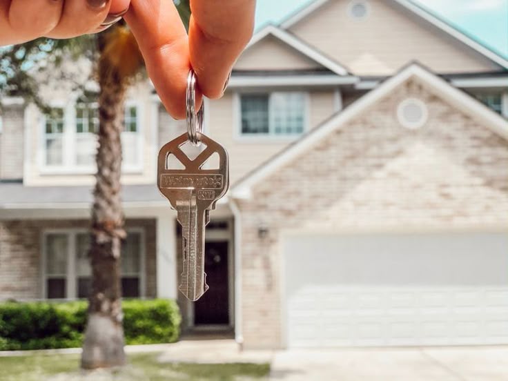 _ Hand holding a key in front of a new brick house, representing homeownership and real estate success. | Sky Rye Design Hand holding a key in front of a new brick house, representing homeownership and real estate success.