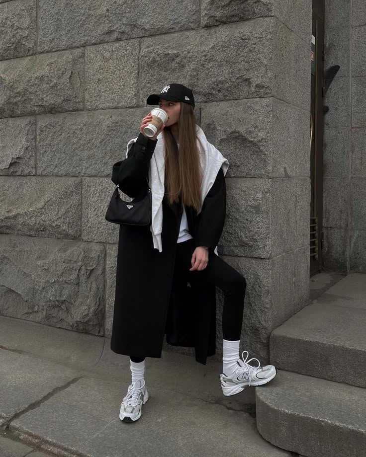 Stylish woman in streetwear, sipping coffee by a stone wall; black coat, sneakers, and cap outfit. Modern urban fashion.