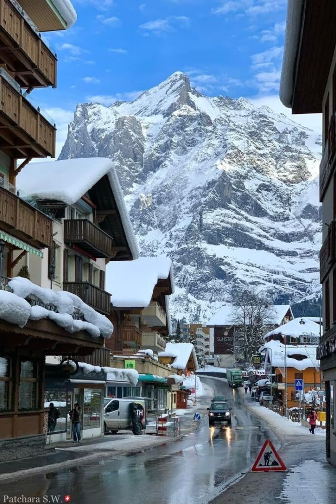 Snow-covered alpine village street with wooden chalets, cars, and a stunning mountain backdrop under a clear blue sky.