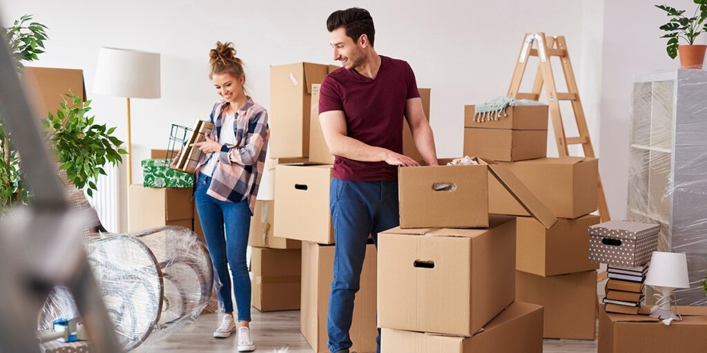 Couple happily packing boxes in a bright room, preparing for a move. Surrounded by plants, a ladder, and home decor items.