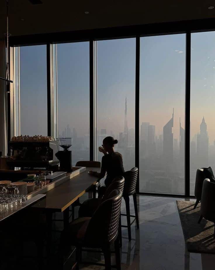 zfgndgb Person enjoying coffee in a modern cafe with a city skyline view through large windows. | Sky Rye Design Person enjoying coffee in a modern cafe with a city skyline view through large windows.