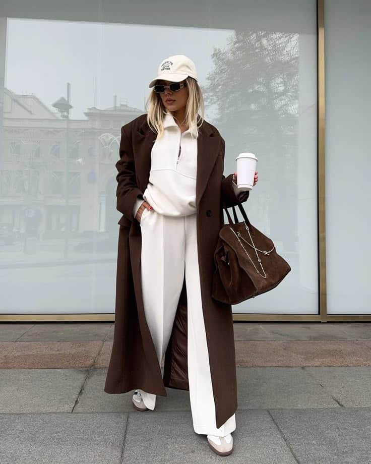 Fashionable woman in a brown coat and white outfit holds coffee and bag, posing stylishly outside a glass building.