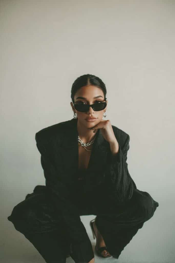 Fashionable woman in black suit and sunglasses, crouching stylishly indoors.