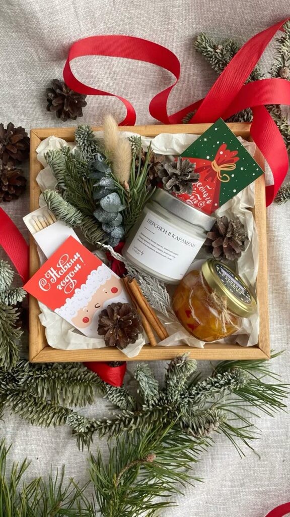 Новогодний подарок Festive holiday gift box with pinecones, greenery, candles, and treats, adorned with red ribbon and Christmas cards on a cozy background. | Sky Rye Design Festive holiday gift box with pinecones, greenery, candles, and treats, adorned with red ribbon and Christmas cards on a cozy background.