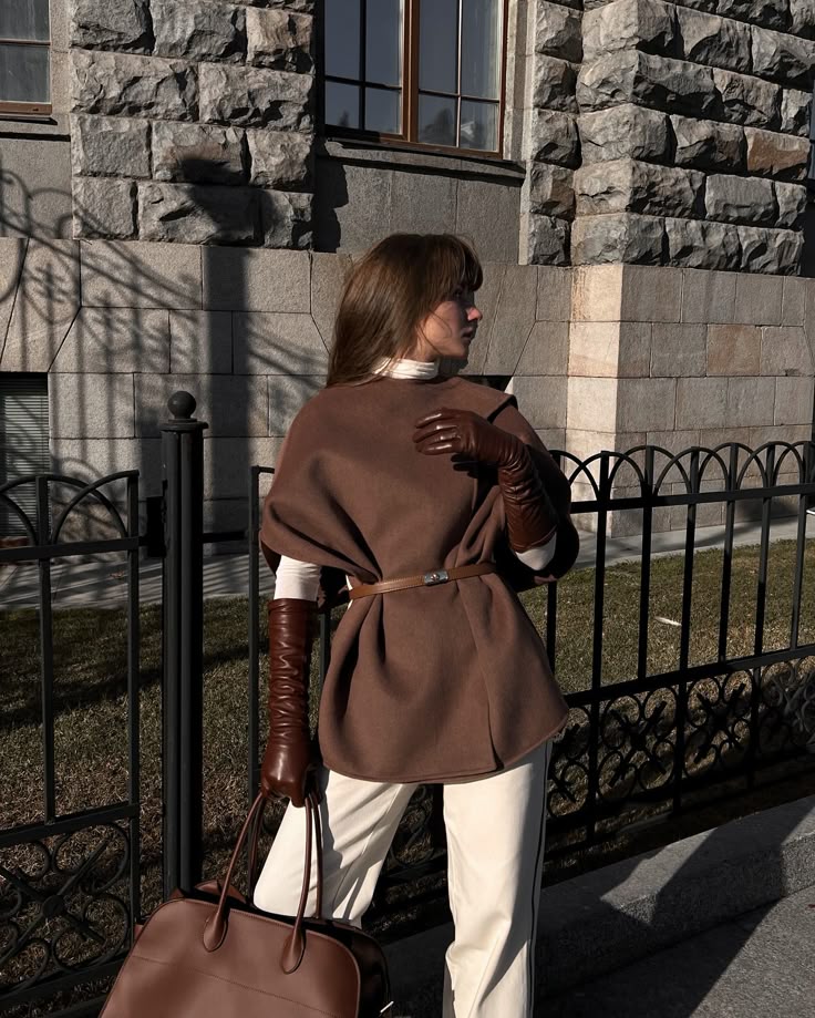 Fashionable woman in brown cape, gloves, and bag, poses with a stone building backdrop. Stylish fall outfit inspiration.
