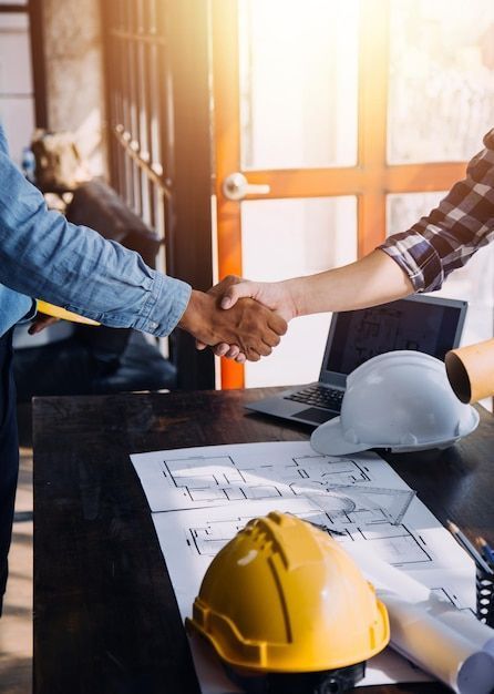 паимфуАЫ Two people shaking hands over a table with construction helmets, blueprints, and a laptop. | Sky Rye Design Two people shaking hands over a table with construction helmets, blueprints, and a laptop.