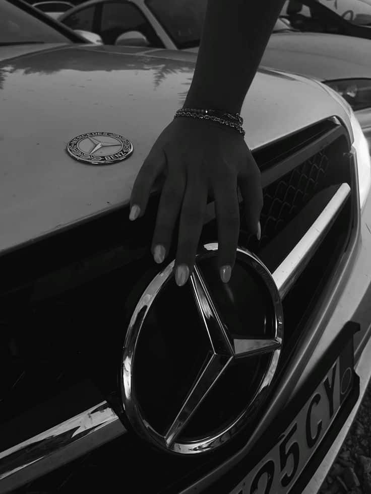яыпаивя Hand resting on a Mercedes-Benz emblem on a car grille, showcasing luxury and style. | Sky Rye Design Hand resting on a Mercedes-Benz emblem on a car grille, showcasing luxury and style.