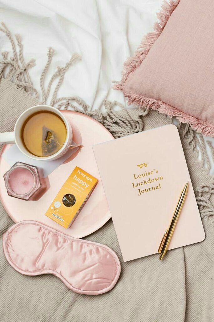 Cozy setup with tea, journal labeled Louise's Lockdown Journal, pink eye mask, and pillow on a soft blanket for relaxation.