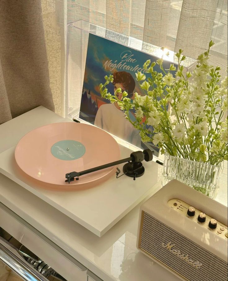 ᑉ³ Turntable playing pastel pink vinyl record, with album cover and flowers in vase, near a retro speaker on a sunny windowsill. | Sky Rye Design Turntable playing pastel pink vinyl record, with album cover and flowers in vase, near a retro speaker on a sunny windowsill.