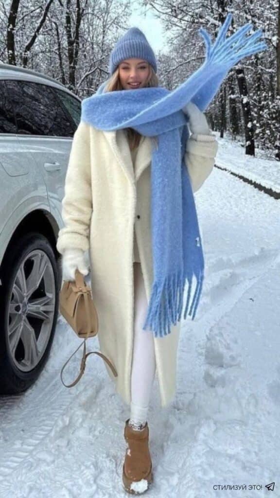 Woman in winter coat and blue scarf walking in snowy park, smiling with brown boots and handbag, beside a car.