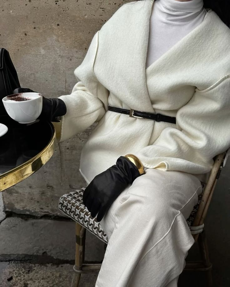 Stylish person in a white coat and gloves enjoys a coffee at a chic café, showcasing elegant winter fashion.