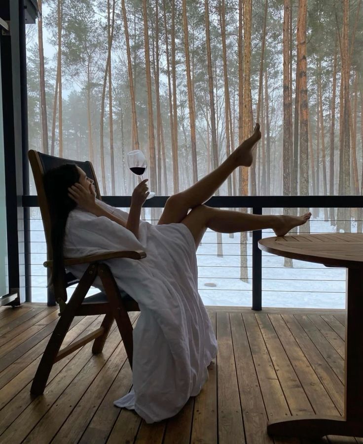 Woman relaxing on a porch in a robe, holding a glass of red wine, with a snowy forest view in the background.