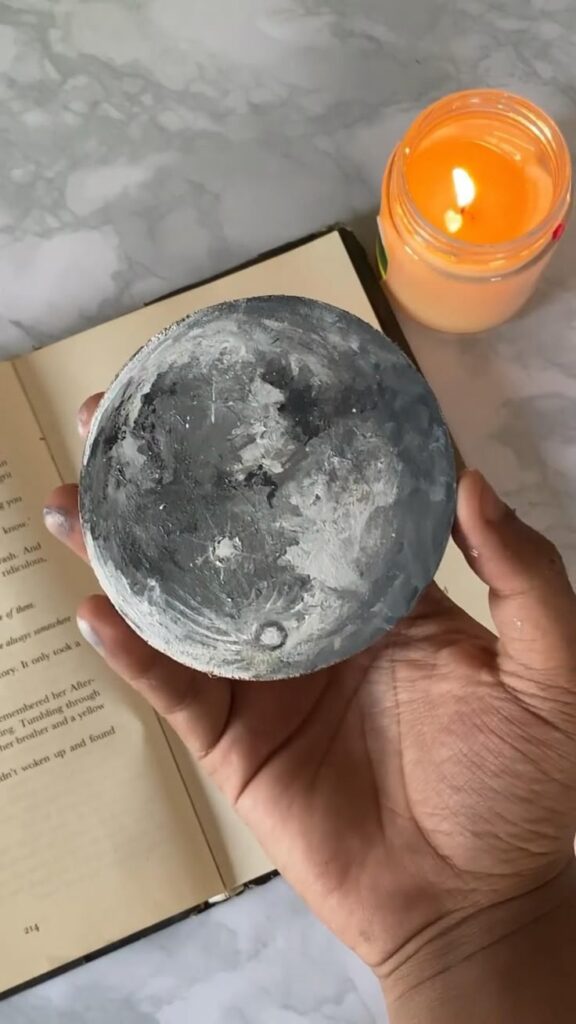 Hand holding moon-shaped art piece above open book, with lit candle nearby on marble surface. Cozy and creative atmosphere.