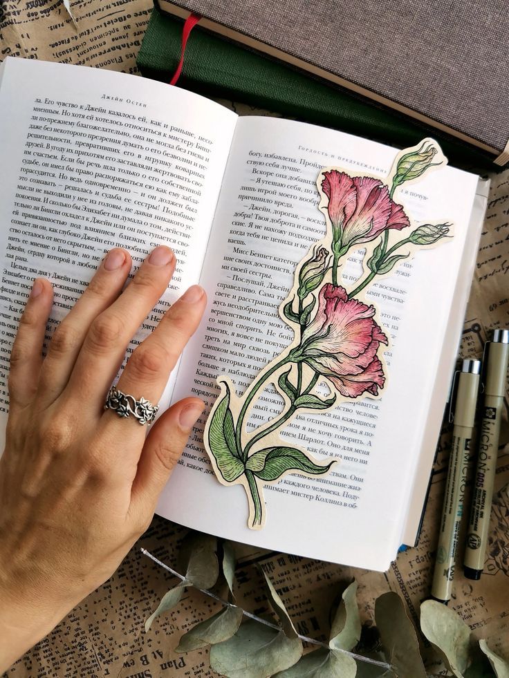 🌷💜🩷🧸🤎🦋🧡❤️ Open book with hand holding a flower bookmark, surrounded by pens and leaves on a newspaper background. Cozy reading scene. | Sky Rye Design Open book with hand holding a flower bookmark, surrounded by pens and leaves on a newspaper background. Cozy reading scene.