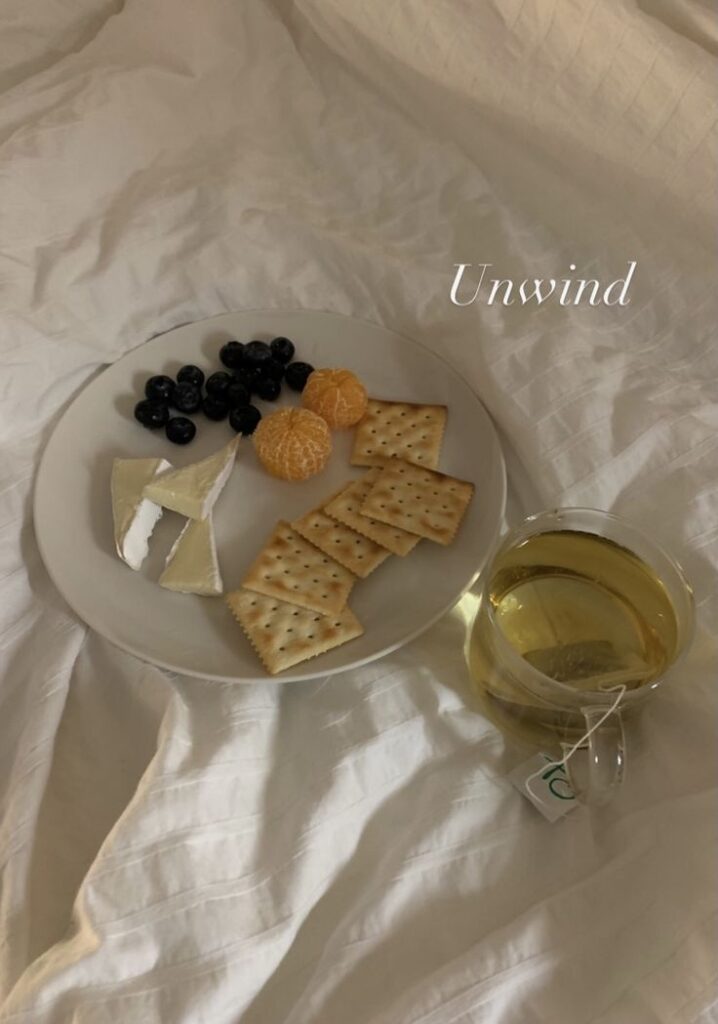 Plate with crackers, Brie cheese, blueberries, mandarins, and a cup of green tea on a white bedspread, text Unwind in the image.