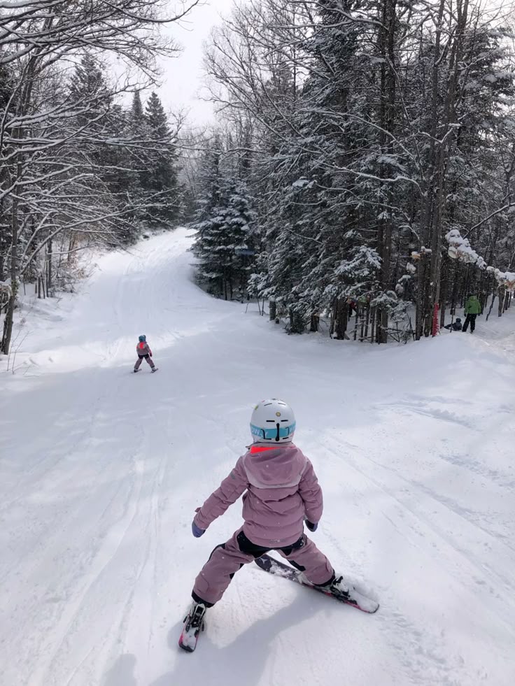 Children skiing on a snowy forest trail, dressed in pink winter gear and helmets, enjoying a fun winter adventure.