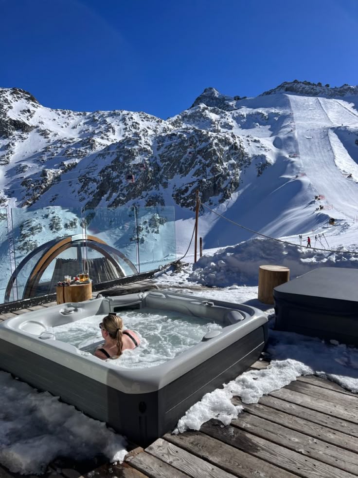 Person relaxing in an outdoor hot tub with a snowy mountain backdrop, under a clear blue sky. Perfect winter retreat setting.