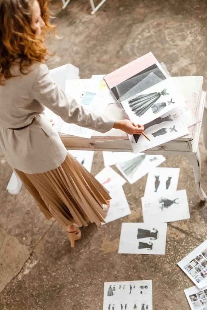 Швейный блог Fashion designer reviews dress sketches on a table, surrounded by design sheets, in a creative studio setting. | Sky Rye Design Fashion designer reviews dress sketches on a table, surrounded by design sheets, in a creative studio setting.