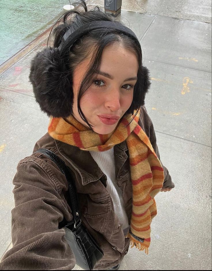 ♡⋆_ Woman wearing earmuffs and striped scarf, posing outdoors on a rainy day. Fashionable winter street style. | Sky Rye Design Woman wearing earmuffs and striped scarf, posing outdoors on a rainy day. Fashionable winter street style.