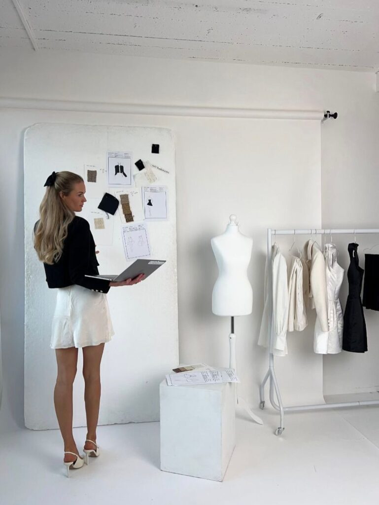 Fashion designer reviewing sketches in a modern studio with mood board, mannequin, and clothing rack. Creative workspace concept.