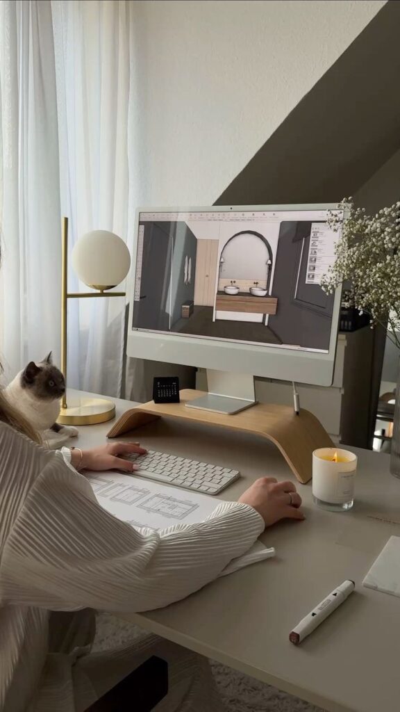 Interior designer working on a 3D model on a computer with a cat nearby, in a cozy workspace with a candle and flowers.