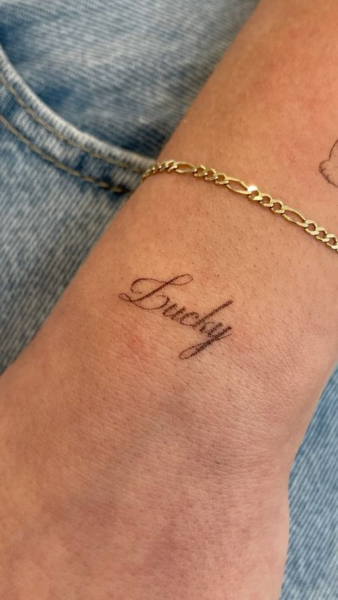 Delicate Lucky script tattoo on wrist, adorned with a gold bracelet against a denim background.