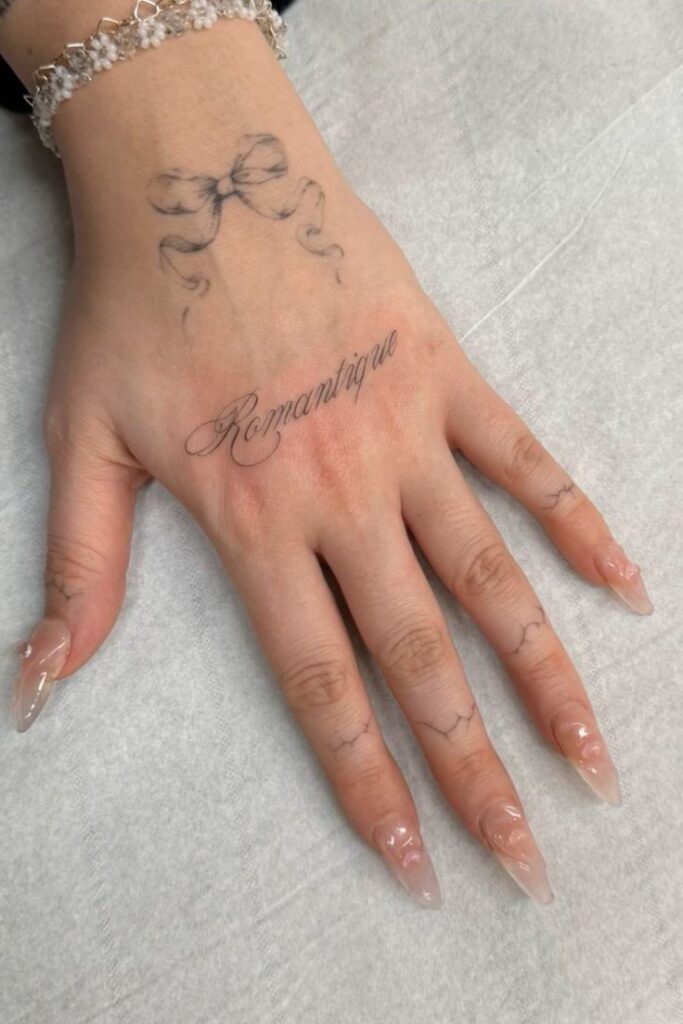 Close-up of a hand with Romantique tattoo and bow, showcasing elegant long nails and a beaded bracelet on a soft background.