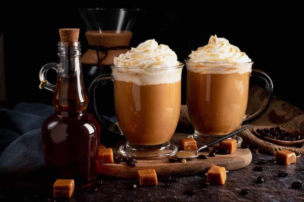 Two caramel lattes topped with whipped cream, surrounded by coffee beans and caramel squares, with a bottle of syrup on the table.