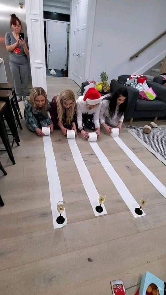 Four women participate in a festive toilet paper race game indoors, with a fifth woman capturing the moment on her phone.