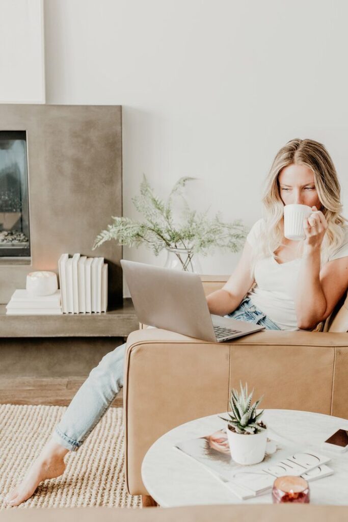 Woman sipping coffee while working on laptop in cozy living room, with plant and books on table, beige couch, modern decor.