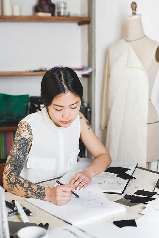 _ 1 Fashion designer sketching in a studio, surrounded by fabric samples and sewing tools, showcasing creativity and design process. | Sky Rye Design Fashion designer sketching in a studio, surrounded by fabric samples and sewing tools, showcasing creativity and design process.
