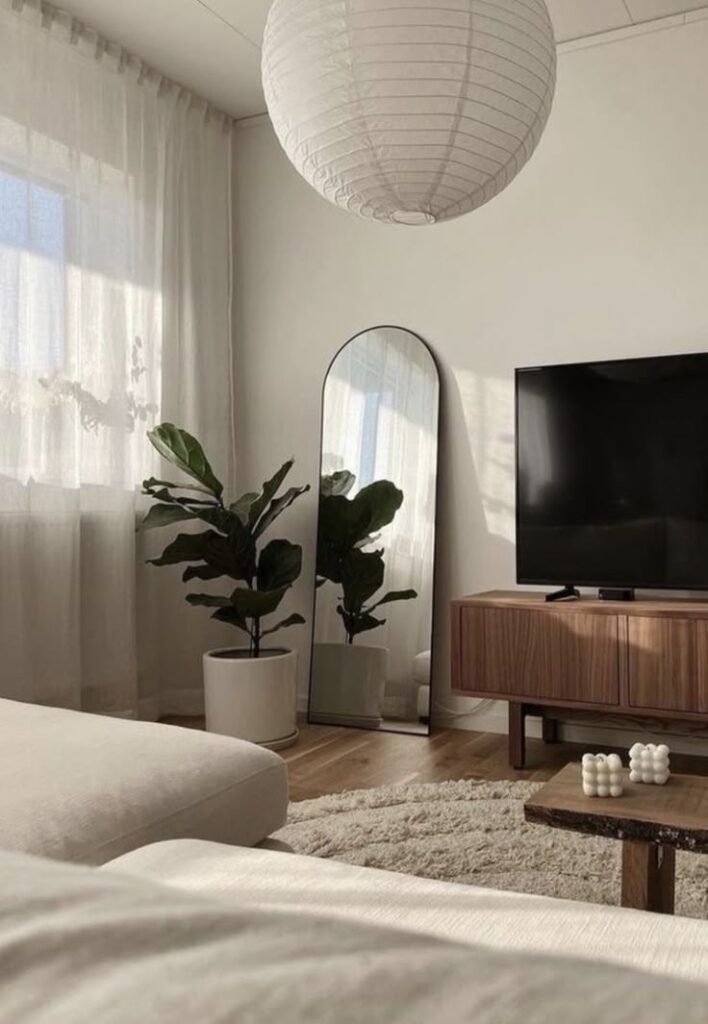 Modern living room with large mirror, potted plant, TV on a wooden stand, cozy beige sofa, and a paper lantern light fixture.