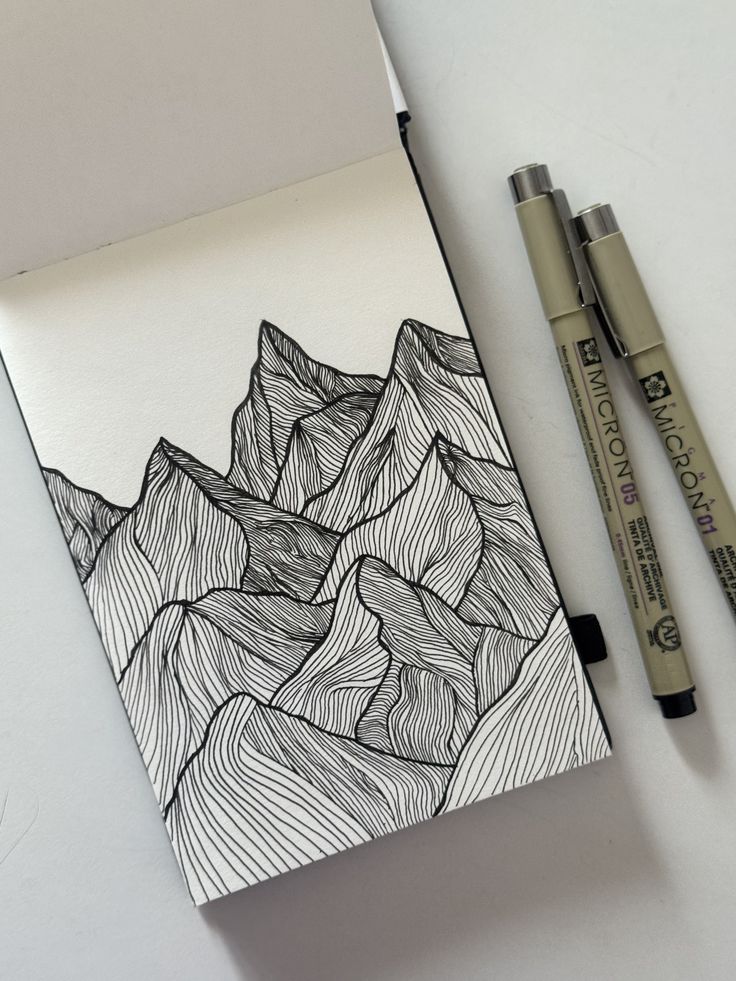 Line art mountain sketch in a notebook with Micron pens beside it, showcasing intricate patterns and creativity.