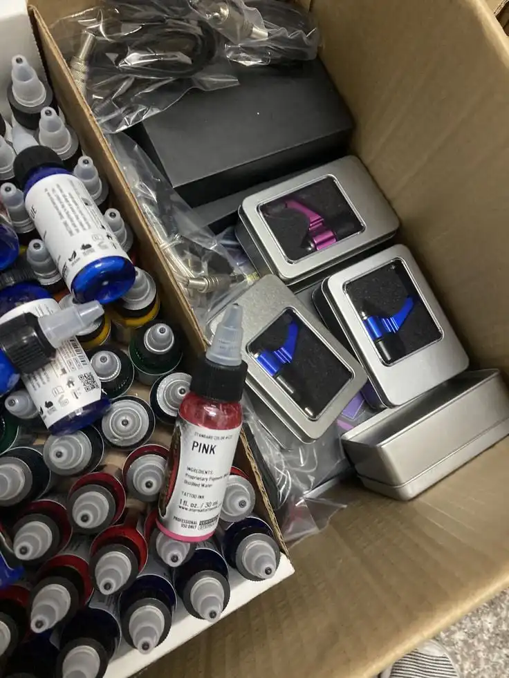 Box of colorful tattoo ink bottles and tattoo machine parts, perfect for professional tattoo artists and enthusiasts looking for supplies.