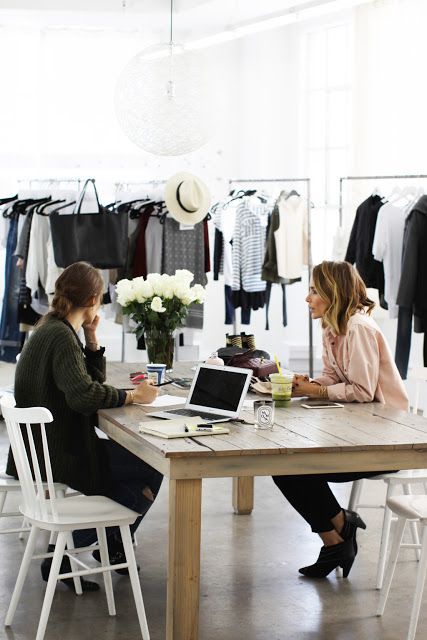 _ 12 Two women working at a fashion studio table with laptops, notebooks, and racks of clothes in the background. | Sky Rye Design Two women working at a fashion studio table with laptops, notebooks, and racks of clothes in the background.