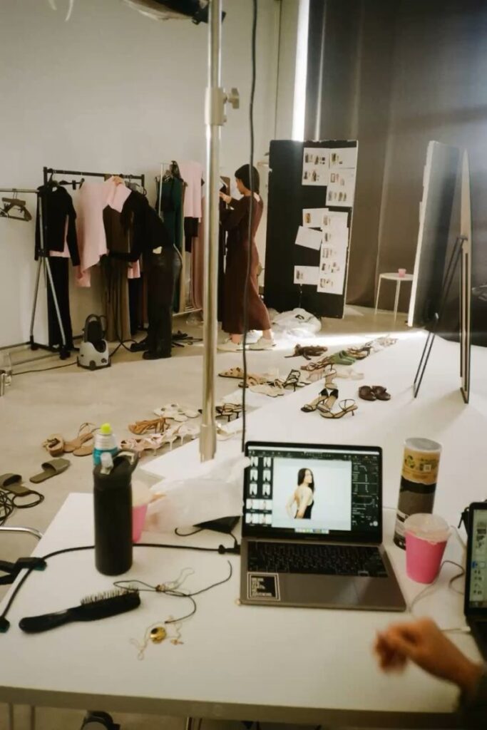 Fashion photoshoot setup with clothing racks, laptop, and accessories in a studio. Models and stylists prepare in a creative workspace.