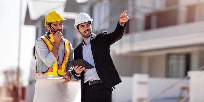12022024121329Blog 3 Two construction professionals discuss a project on-site, wearing hard hats. One holds blueprints while the other points with a tablet in hand. | Sky Rye Design Two construction professionals discuss a project on-site, wearing hard hats. One holds blueprints while the other points with a tablet in hand.