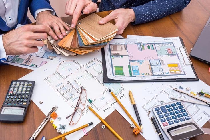 ArchitectsDiscussWhichTeamToChooseTheColorOfThe Architects reviewing floor plans with calculators and swatches on a desk, planning interior design and materials for a construction project. | Sky Rye Design Architects reviewing floor plans with calculators and swatches on a desk, planning interior design and materials for a construction project.