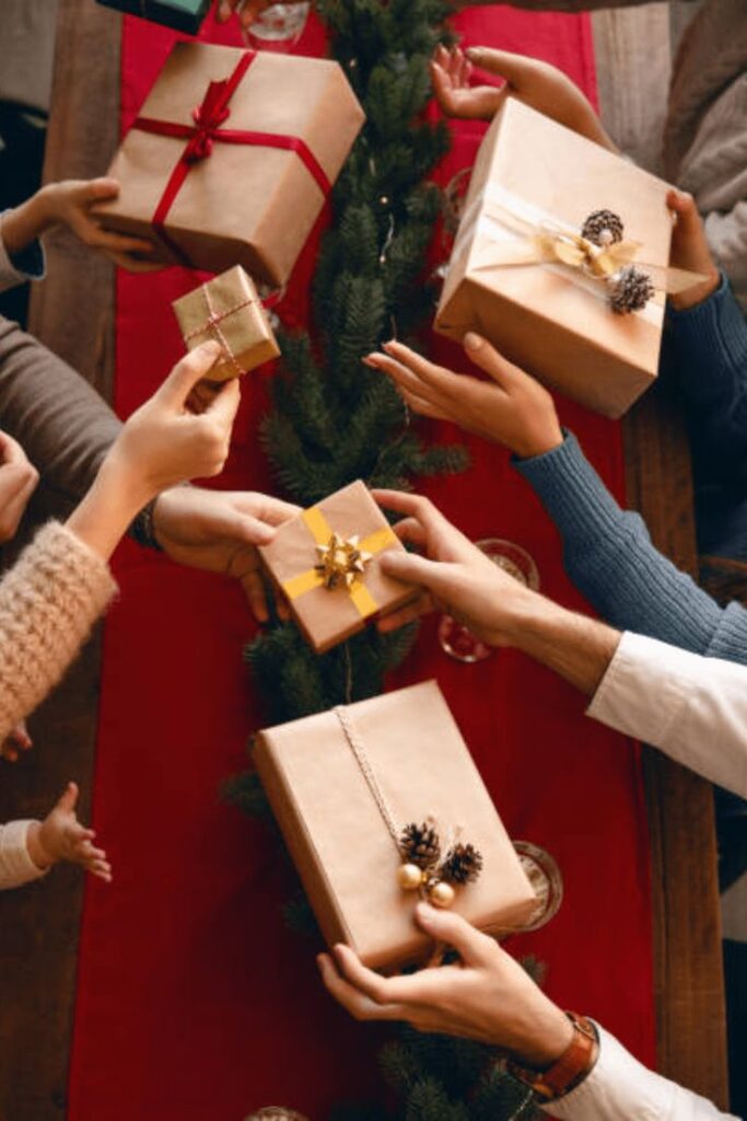 People exchanging wrapped gifts around a festive table with a red runner and greenery, signifying holiday celebration and sharing.