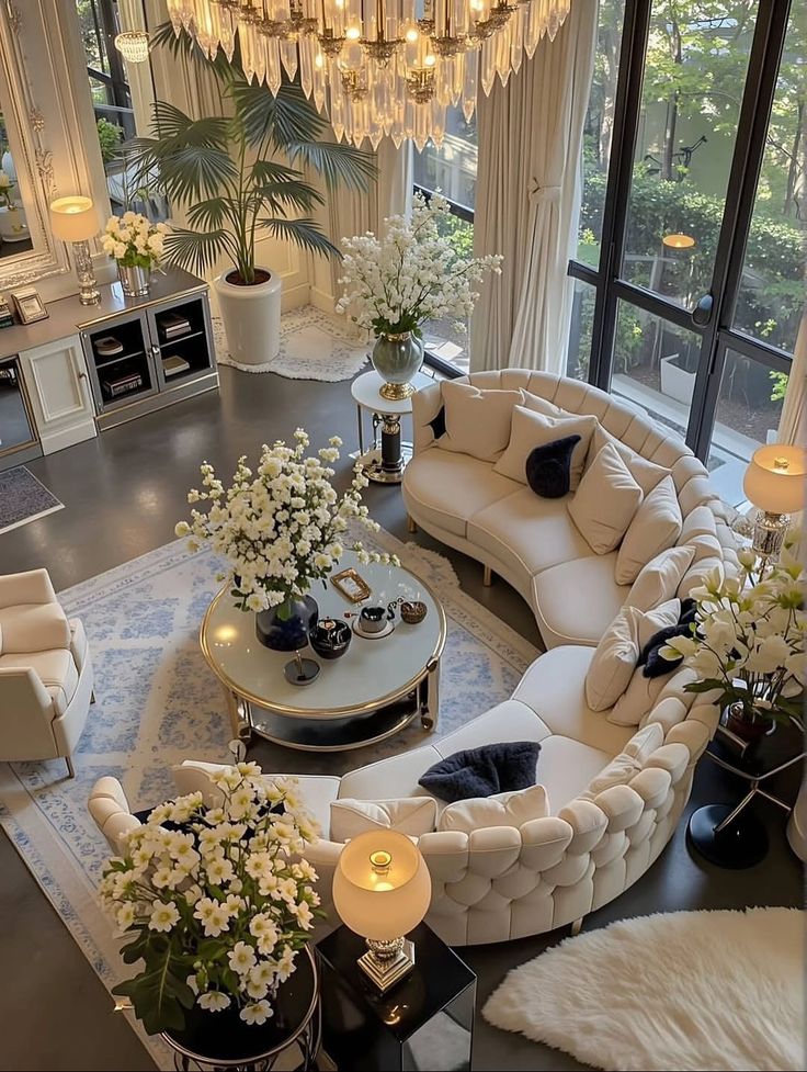 13 Luxe Yet Cozy White and Gold Living Room Ideas That Wow Luxurious living room with elegant chandelier, curved sofas, floral decor, and large windows offering a view of greenery outside. | Sky Rye Design Luxurious living room with elegant chandelier, curved sofas, floral decor, and large windows offering a view of greenery outside.
