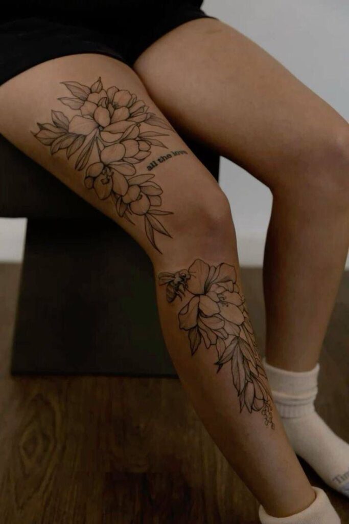 _ 14 Thigh tattoo featuring intricate floral design and bee, with text all the love, on a person sitting with crossed legs. | Sky Rye Design Thigh tattoo featuring intricate floral design and bee, with text all the love, on a person sitting with crossed legs.