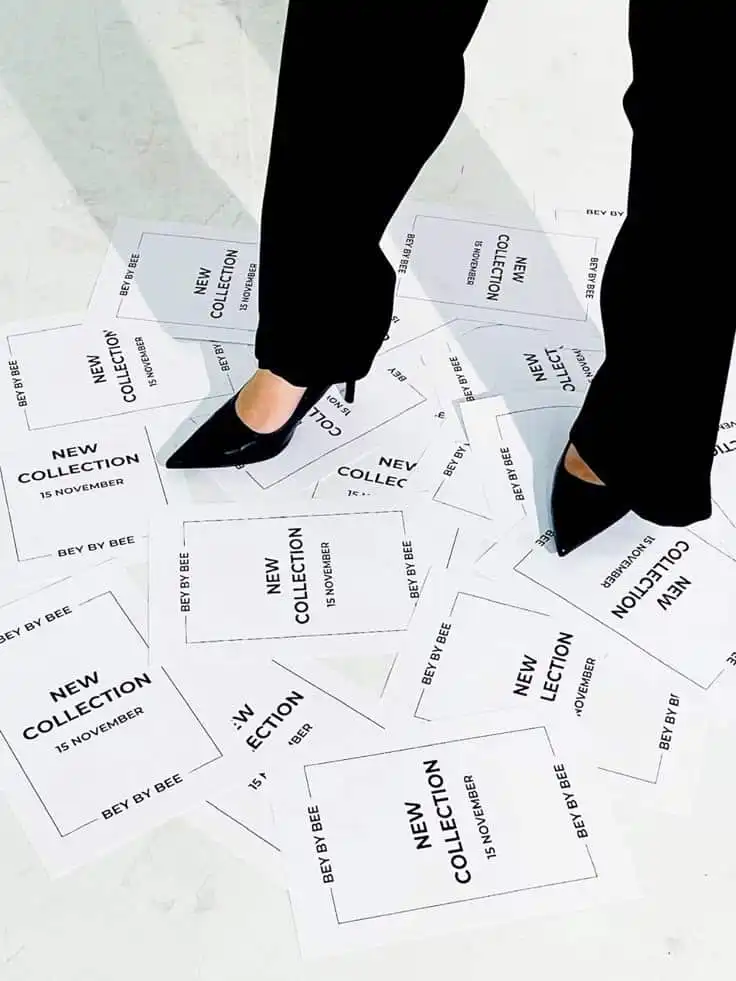 Person standing on flyers announcing New Collection launch by Bey By Bee on November 15, wearing stylish black shoes.