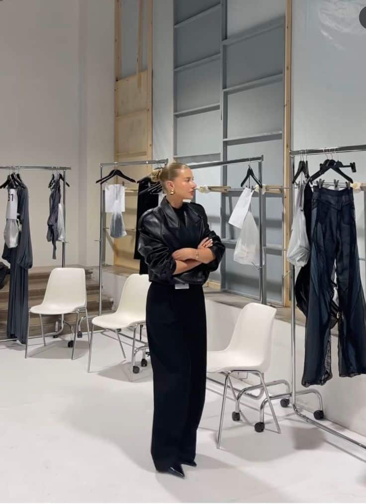 _ 15 Fashion stylist in a black outfit stands in a modern studio, surrounded by clothing racks and white chairs. | Sky Rye Design Fashion stylist in a black outfit stands in a modern studio, surrounded by clothing racks and white chairs.
